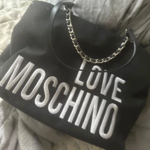 Svart handväska från Love Moschino - Stor svart handväska från Love Moschino med tydlig vit logotyp framtill. Väskan har kedjedetaljer på axelremmen och dragkedja inuti. Perfekt för dig som vill ha en snygg och rymlig accessoar.