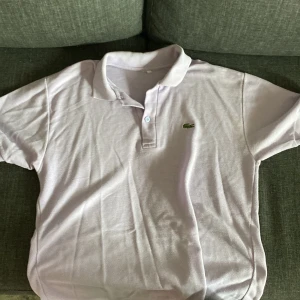 Ljuslila pikétröja från Lacoste - Säljer en klassisk ljuslila pikétröja från Lacoste med den ikoniska gröna krokodilloggan på bröstet. Tröjan har korta ärmar, krage och knäppning med två knappar framtill. Perfekt för en stilren och sportig look.
