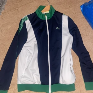 Blåvit track jacket från Lacoste - Säljer en snygg track jacket från Lacoste i blått och vitt med gröna detaljer. Jackan har dragkedja, hög krage och logga på bröstet. Perfekt för en sportig look och har långa ärmar.