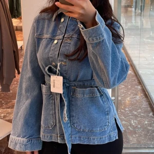 Jeansjacka med bälte - Slutsåld blå jeansjacka med knappar och knytskärp i midjan från Åhléns (Carin Wester). Jackan har stora fickor framtill och lång ärm. Smickrande och perfekt för vår och sommar. Lånade bilder. Nyskick. 