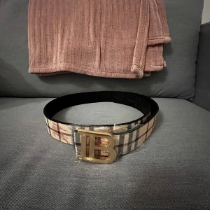 Rutmönstrat bälte med guldspänne - Säljer ett snyggt bälte med klassiskt rutmönster i beige, svart. Bältet har ett stort guldigt B-spänne och insidan är svart. Perfekt accessoar för att lyfta din outfit. Du kan ha båda svart och beige färg på de