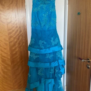 Turkos balklänning med broderier och paljetter - Säljer en turkos långklänning med vackra blommiga broderier och paljettdetaljer. Klänningen har ett axelband där man kan sätta dom blommorna på, volangdetaljer och snörning i ryggen för justerbar passform. Perfekt för bal eller festliga tillfällen.