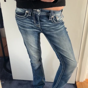 Blå jeans med broderade bakfickor - Säljer ett par blå jeans med snygga slitningar och vita kontrastsömmar. Bakfickorna har detaljerade broderier i grått och vitt. Jeansen har låg midja och klassisk femficksdesign. Perfekta för dig som gillar unika detaljer!