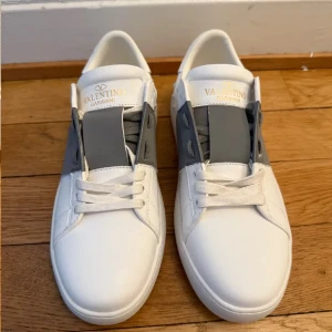 Valentino Garavani vita sneakers med grå detalj - Säljer ett par vita sneakers från Valentino Garavani med bred grå rem över snörningen och ikoniska nitar baktill på sulan. Skorna har rund tå och platt sula, perfekt för en stilren look.