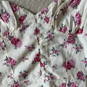 Vit blommig klänning med knappar från Zara - Supersöt klänning från Zara med smala axelband och blommigt mönster i rosa och rött. Klänningen har knappar framtill, liten slits och fina broderade detaljer. Perfekt för sommarens alla tillfällen! Klänningen är oanvänd, för stor för mig. 