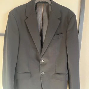 Svart kavaj från Zara Slim Tailoring - Säljer en svart kavaj från Zara med slim passform. Kavajen har två knappar framtill, klassiska slag och fickor med lock. Endast använd på min student. Strlk s-m. Hör av vid frågor eller fler bilder!