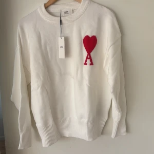 Vit stickad tröja från Ami - Vit stickad tröja från Ami med rund halsringning och röd broderad hjärtdetalj med bokstaven A på bröstet. Tröjan har långa ärmar och breda ribbade muddar vid ärmslut och nederkant. Perfekt för en stilren och trendig look.
