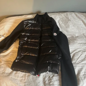 Svart pufferjacka - Säljer en svart pufferjacka från Moncler med glansig vadderad front och stickade ärmar. Jackan har dragkedja och Moncler-logga på ena ärmen. Perfekt för kyliga dagar och snygg till streetwear. Nfc tagg fungerar 