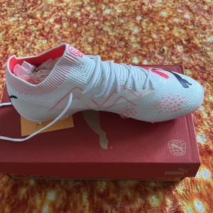 Vita fotbollsskor från Puma - Säljer ett par vita fotbollsskor från Puma med röda och svarta detaljer. Skorna har snörning och en stickad, strumpliknande ovandel för extra komfort och passform. Perfekta för dig som vill sticka ut på planen!