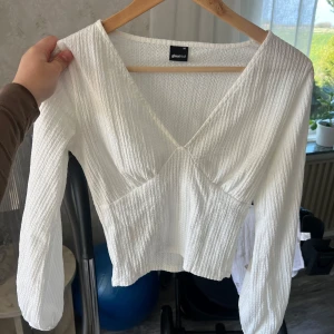 Vit v-ringad topp från Ginatricot - Säljer en vit långärmad topp med v-ringning och lätt ribbad struktur. Toppen har en figurnära passform och är tillverkad i ett mjukt syntetmaterial. Perfekt att styla med jeans eller kjol.