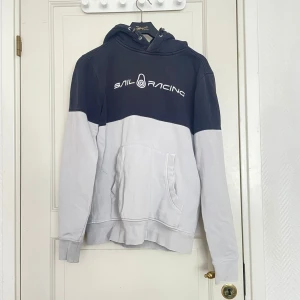 Svart och grå hoodie från Sail Racing - Säljer en snygg hoodie från Sail Racing i svart och ljusgrått. Tröjan har huva med dragsko, stor magficka och logga broderad på bröstet. Perfekt för en sportig och avslappnad stil.