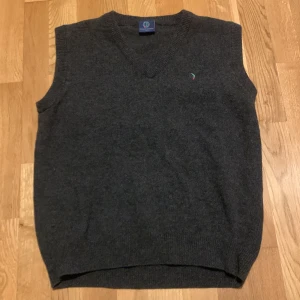 Mörkgrå ullväst från Boomerang - Mörkgrå stickad väst från Boomerang i ull med v-ringad hals. Liten broderad logotyp på bröstet i grönt och rött. Klassisk och enkel design som passar till skjorta eller t-shirt.