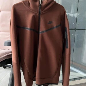 Brun Nike Tech Fleece - Säljer en brun tech fleece från Nike med svarta detaljer och dragkedja framtill. Jackan har huva och en diskret ficka med dragkedja på ärmen. Helt oanvänd och otvättad. Storlek L