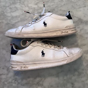 Vita sneakers från Polo Ralph Lauren - Säljer ett par klassiska vita sneakers från Polo Ralph Lauren med blå logga på sidan och mörkblå detalj vid hälen. Skorna har snörning, rund tå och platt sula. Tillverkade i syntet och skinnblandning. Perfekta till vardags för en clean och stilren look. Inga hål. 1250kr nypris men säljer för endast 299kr