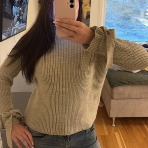 Beige stickad tröja med knytband - Säljer en beige stickad tröja med snygga knytband vid ärmsluten. Tröjan har lång ärm och en rak, något loose passform. Perfekt för dig som gillar stilrena detaljer och mjuka material.