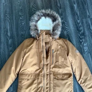 Beige parkajacka med fuskpäls från Name It - Säljer en beige parkajacka från Name It med huva och fuskpäls. Jackan har dragkedja, flera fickor och en mysig krage. Perfekt för kalla dagar och riktigt snygg stil! 🧥✨
