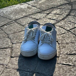 Vita sneakers med gröna detaljer - Säljer ett par klassiska vita sneakers med gröna detaljer på hälen. Skorna har snörning och en platt sula, perfekt för en stilren och enkel look. Passar till det mesta och är riktigt bekväma att ha på sig. Storlek 40. Kan förhandlas/ställ gärna frågor.
