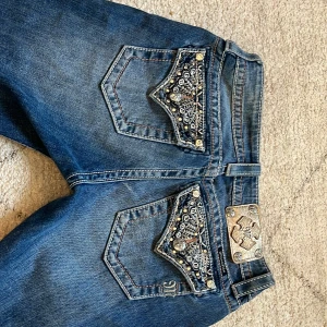 Miss Me Jeans - Säljer ett par blå jeans med snygga detaljerade bakfickor med paljetter och nitar. Jeansen har bootcut-modell och klassisk jeanslook. Perfekta för dig som vill sticka ut lite extra med dina byxor.