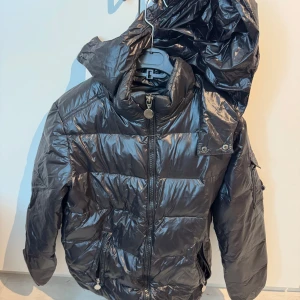 Svart dunjacka från Moncler(fejk) - Säljer en svart glansig dunjacka som liknar Moncler med huva och dragkedja framtill. Jackan har Moncler-logga på ärmen, praktiska fickor och hög krage. Perfekt för kyliga dagar när du vill hålla stilen.