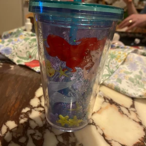 Disney Ariel plastmugg med sugrör - Säljer en genomskinlig plastmugg med blått lock och sugrör. Motivet är Ariel från Den lilla sjöjungfrun, tillsammans med Flounder och undervattensdetaljer i färgglada nyanser. Perfekt för kalla drycker och har ett lekfullt, färgstarkt utseende.
