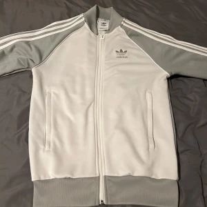 Vit och grå Adidas track jacket - Säljer en klassisk Adidas track jacket i vitt och ljusgrått med dragkedja framtill. Jackan har de ikoniska tre ränderna längs ärmarna och två fickor med dragkedja. Perfekt för en sportig och avslappnad stil.