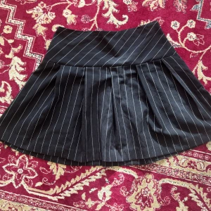 Svart kortkjol med vita ränder - Säljer en svart kortkjol med vita tunna ränder i klassiskt kostymtyg. Kjolen har veck framtill och dragkedja bak. Perfekt för en stilren och trendig look.