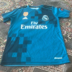 Real Madrid bortatröja Ronaldo 7 - Snygg blå Real Madrid bortatröja från Adidas med Ronaldo 7 på ryggen. Tröjan har korta ärmar, pixelmönster i mörkblått och klubbmärke samt sponsortryck på bröstet. Tillverkad i lätt och ventilerande material med climacool-teknologi.