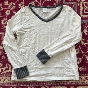 Vit långärmad tröja med randiga detaljer från Stick & Stay - Vit långärmad tröja från Stick & Stay med svarta och vita ränder på v-ringad krage och muddar. Mjuk och skön bomullskvalitet, perfekt för en avslappnad stil. Klassisk passform och stilren design.