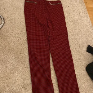 Röda rutiga kostymbyxor från MNG - Säljer ett par röda kostymbyxor från MNG med svart rutigt mönster och dragkedjedetaljer vid fickorna. Byxorna har en klassisk passform och är perfekta för dig som vill sticka ut lite extra.