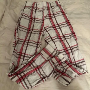 Rutig pyjamasbyxa i bomull - Säljer ett par mjuka och bekväma pyjamasbyxor i vitt med rött och lila rutmönster. Byxorna har resår i midjan och är perfekta för att chilla hemma eller sova i. Klassisk och avslappnad stil.