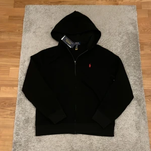 Svart hoodie från Polo Ralph Lauren - Säljer en svart hoodie från Polo Ralph Lauren med dragkedja och huva. Klassisk design med röd broderad logga på bröstet. Perfekt för en avslappnad och stilren look. Passar som S. Helt ny med prislapp. Og pris 1499 vårt pris 599. Returrätten gick ut.