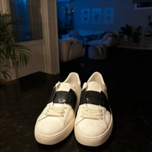 Valentino Garavani Open sneakers i vitt och svart - Säljer ett par snygga Valentino Garavani Open sneakers i vitt skinn med svart detalj över mitten. Skorna har platt sula, rund tå och ikoniska nitar på hälen. Perfekta för dig som gillar exklusiva och stilrena sneakers.