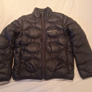 Svart dunjacka från Peak Performance - Säljer en svart dunjacka från Peak Performance med hög krage och dragkedja framtill. Jackan har quiltad design och logga på bröstet samt ärmen. Perfekt för kyliga dagar och enkel att matcha med det mesta.