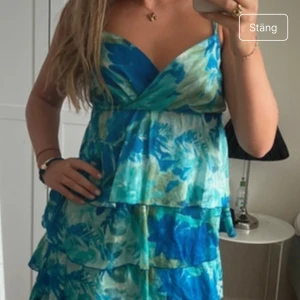 Blågrön blommig volangklänning - Säljer en kort klänning med blommigt mönster i blått, grönt och vitt. Klänningen har volanger i flera lager och omlott över bysten. Perfekt för sommarens alla tillfällen!