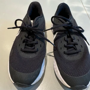 Svarta Nike sneakers med vit sula - Säljer ett par svarta Nike sneakers med vit sula och vit swoosh på sidan. Skorna har snörning och en sportig design, perfekta för träning eller vardagsbruk. Ovandelen är i mesh-liknande syntetmaterial för bra andningsförmåga. Helt nya aldrig använda. Strl 37.5