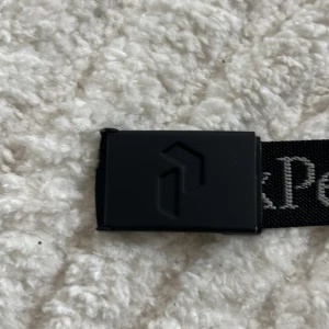 Svart bälte från Peak Performance /104 CM  - Säljer ett svart bälte från Peak Performance med tydlig logotyp och text längs bandet. Bältet har ett svart spänne i metall och är gjort i slitstarkt tygmaterial. Perfekt  för en stilren look🤩 Odinarie pris 349kr mitt pris 99kr 
