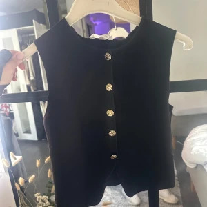 Svart väst med guldknappar - Stilren svart väst med dekorativa guldknappar framtill. Västen har en rak passform och är ärmlös, perfekt att styla över en skjorta eller t-shirt för en trendig look. Endast använd en gång. 