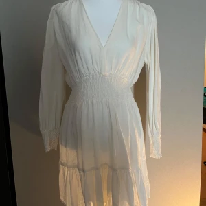 Vit klänning  - Säljer en söt vit klänning med v-ringning och långa ärmar. Klänningen har smock i midjan och vid ärmsluten samt en lätt volang nertill. Från zara