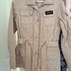 Beige jacka från Barbour - Snygg beige quiltad jacka från Barbour med bälte i midjan och flera fickor framtill. Jackan har lång ärm, krage och knappar samt dragkedja. Perfekt för övergångssäsonger och ger en klassisk look.
