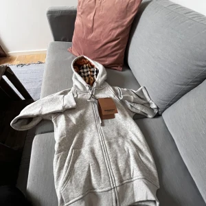 Grå hoodie från välkänt märke - Säljer en grå hoodie  med dragkedja och klassiskt rutigt foder i huvan. Tröjan har justerbar dragsko vid huvan och är perfekt för en avslappnad stil. Tröjan passar liten S ca 165-170