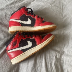 Nike Air Jordan 1 Mid svart/röd/vit - Säljer ett par Nike Air Jordan 1 Mid med svart bas, röda detaljer och vit swoosh. Skorna har klassisk rund tå, snörning och platt sula. Perfekta för dig som gillar streetwear och sneakers med ikonisk design.