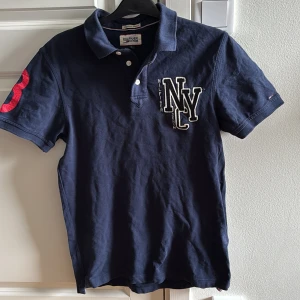 Hilfiger Denim Piké - Säljer en mörkblå pikétröja från Hilfiger Denim med broderad NYC-logga på bröstet och röd siffra på ärmen. Klassisk krage och knäppning framtill. Perfekt för en avslappnad och stilren look.
