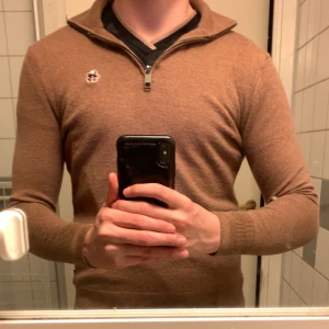 Brun halvzip tröja från Ted baker - Säljer en brun stickad halvzip tröja från Ted baker med liten broderad logga på bröstet. Tröjan har lång ärm, ribbade muddar och krage med dragkedja. Perfekt för lager-på-lager och snygg till jeans.