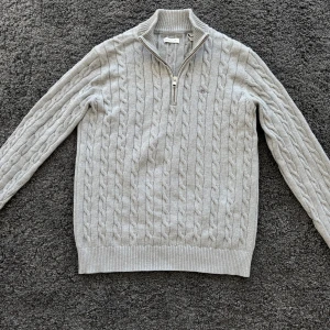 Cotton cable Half zip  - Säljer en ljusgrå stickad tröja från GANT med halv dragkedja och kabelstickat mönster. Tröjan har hög krage och broderad logga på bröstet. Perfekt för kyliga dagar. Sparsamt använd och köpt för en månad sedan på Emporia 