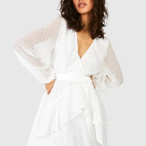 Vit omlottklänning med volang från Boohoo - Superfin vit omlottklänning från Boohoo med volangdetaljer och prickigt mönster. Klänningen har långärmade, transparenta ärmar och markerad midja med knytband. Perfekt för sommarens tillfällen!