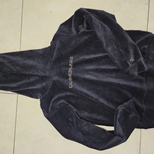 Svart manchesterhoodie Calvin Klein Jeans - Mörk, svart hoodie från Calvin Klein Jeans i mjuk manchester. Tröjan har broderad logga på bröstet och ett litet CK-broderi på ärmen. Klassisk huva och långa ärmar, perfekt för en chill och stilren look.
