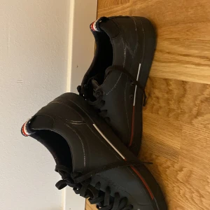 Svarta sneakers från Tommy Hilfiger - Svarta sneakers från Tommy Hilfiger med klassiska röda, vita och blå detaljer. Skorna har snörning, rund tå och coolt mönstrad sula med stadssilhuett. Tillverkade i syntetmaterial med logga på plös och häl.