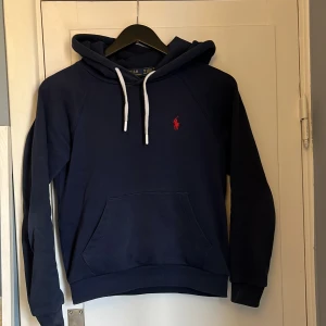 Mörkblå hoodie från Polo Ralph Lauren - Säljer en mörkblå hoodie från Polo Ralph Lauren med klassisk röd logga broderad på bröstet. Tröjan har huva med vita snören och en stor magficka. Perfekt för dig som gillar en enkel men stilren look.