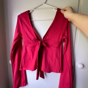 Röd omlottopp med knyt från Nelly - Säljer en snygg röd omlottopp från Nelly i storlek M. Toppen har långa ärmar, djup v-ringning och knyt framtill som ger en trendig look. Tillverkad i mjukt material som känns skönt mot huden. Perfekt för dig som vill sticka ut lite extra.