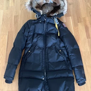 Svart Parajumpers dunjacka med päls - Säljer en svart lång dunjacka från Parajumpers med stor huva fodrad i mjuk päls och snygg fuskpäls runt kanten. Jackan har dragkedja, stora fickor med knappar och den klassiska gula remmen vid halsen. Perfekt för kalla vinterdagar.